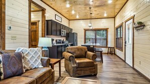 A cozy cabin interior with a wooden ceiling, a dining area, a kitchen, and a living room with leather furniture.