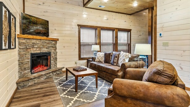 A cozy living room with a stone fireplace, wooden walls, a leather sofa, and a television on a wooden shelf.