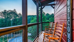 A wooden balcony with a 'WELCOME' sign, wooden chairs, and a view of a forested area.