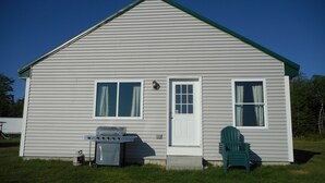 A single-story house with a green roof, two windows, a door, and a barbecue grill in the front yard. - Post and beam cabin with bunk beds (Shirley Mills)