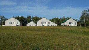 A row of white, single-story houses with green roofs, set in a grassy field with trees in the background. - Post and beam cabin with bunk beds (Shirley Mills)