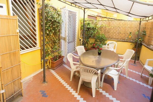A patio with a round table and white plastic chairs, surrounded by yellow walls and potted plants.