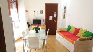 Ein Essbereich mit einem Glastisch, weißen Stühlen und einem kleinen Topfpflanzen. Ein Sofa mit bunten Kissen und ein Fernseher im Hintergrund sind ebenfalls zu sehen.