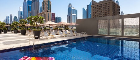 A rooftop pool with lounge chairs and umbrellas, surrounded by city buildings.