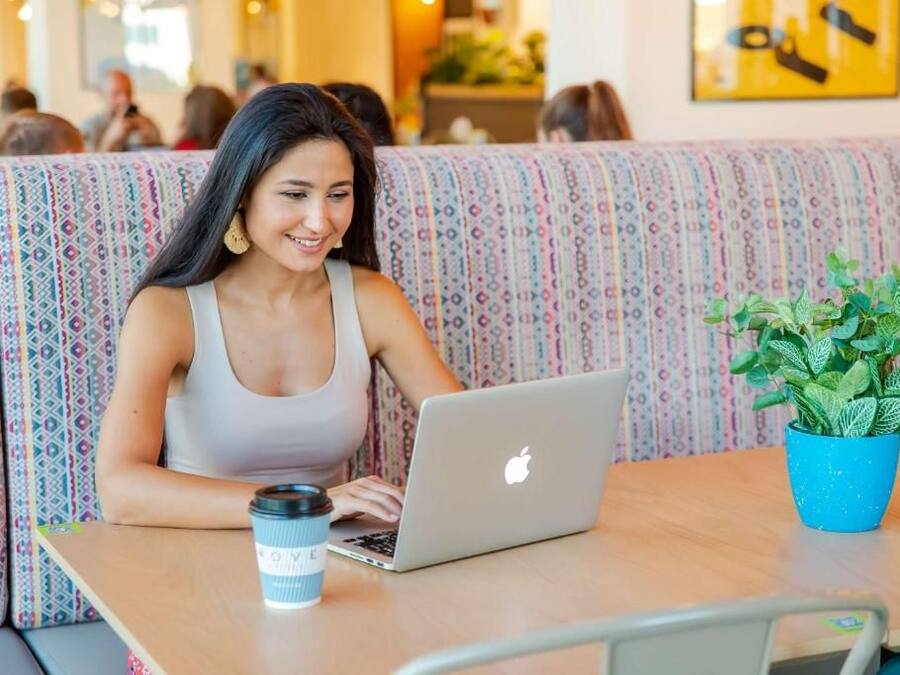 Une personne est assise à une table, devant un ordinateur portable et une tasse de café. En arrière-plan, on aperçoit un canapé à motifs et une plante en pot.