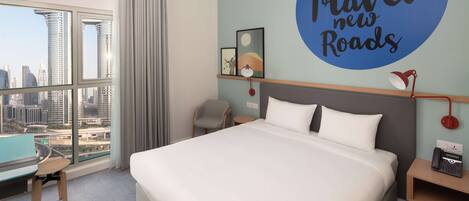 A hotel room with a large bed, a desk, a chair, a city view, and a wall with a motivational quote.