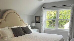 Une chambre avec un grand lit, un ventilateur de plafond, une fenêtre avec des rideaux, une table de chevet et un tableau encadré au mur.