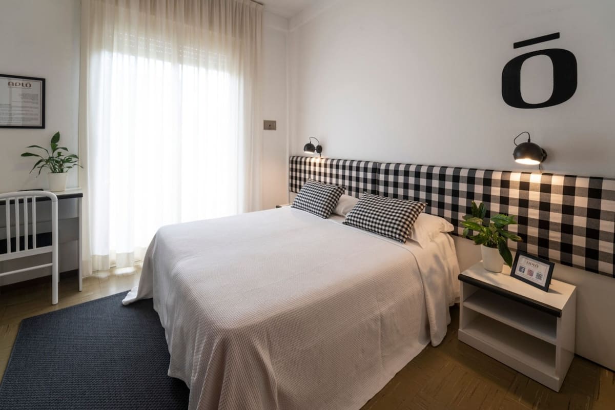 Un dormitorio moderno con una cama grande, cabecera a cuadros, mesitas de noche, una planta en maceta y un aplique de pared.