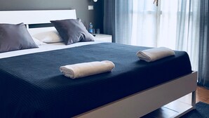 Una habitación de hotel moderna con una cama grande, una mesita de noche con una botella de agua y vasos, y una ventana con cortinas traslúcidas.