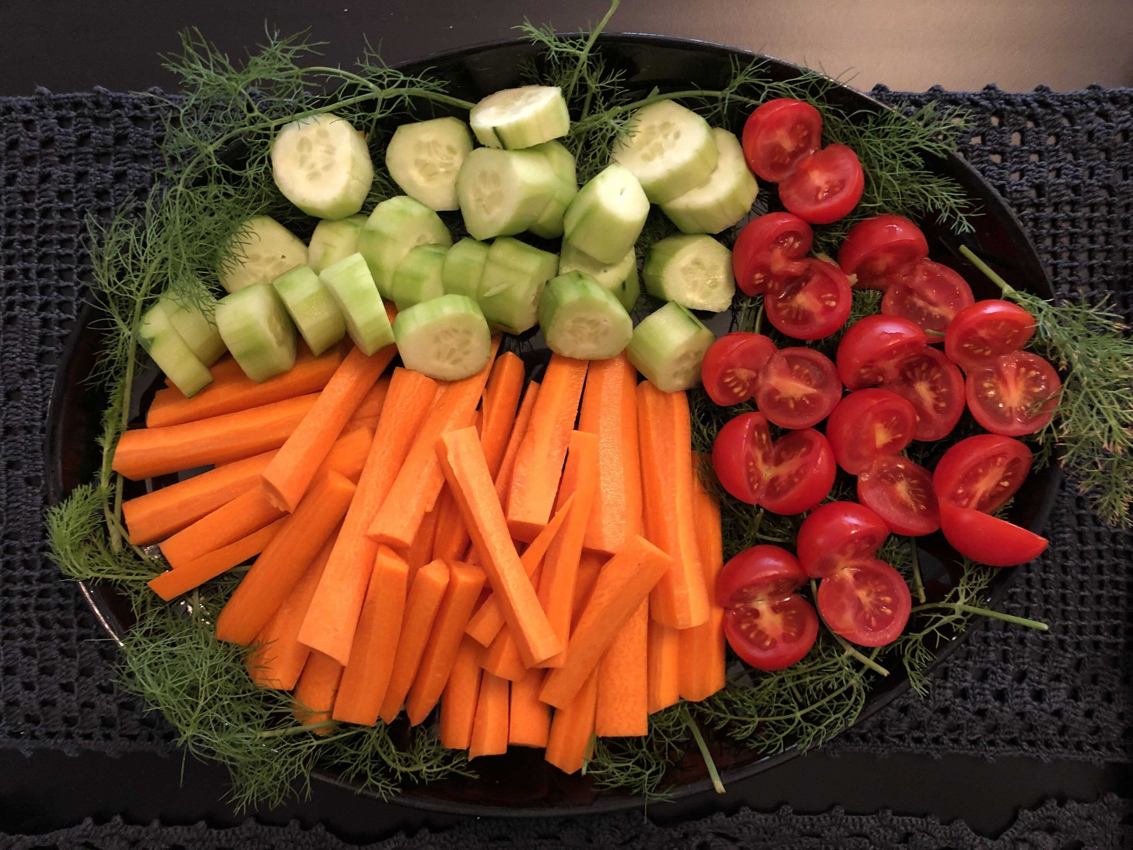Une assiette de légumes coupés en tranches, notamment des concombres, des carottes et des tomates, disposés sur une assiette foncée, elle-même posée sur une nappe foncée à texture marquée.