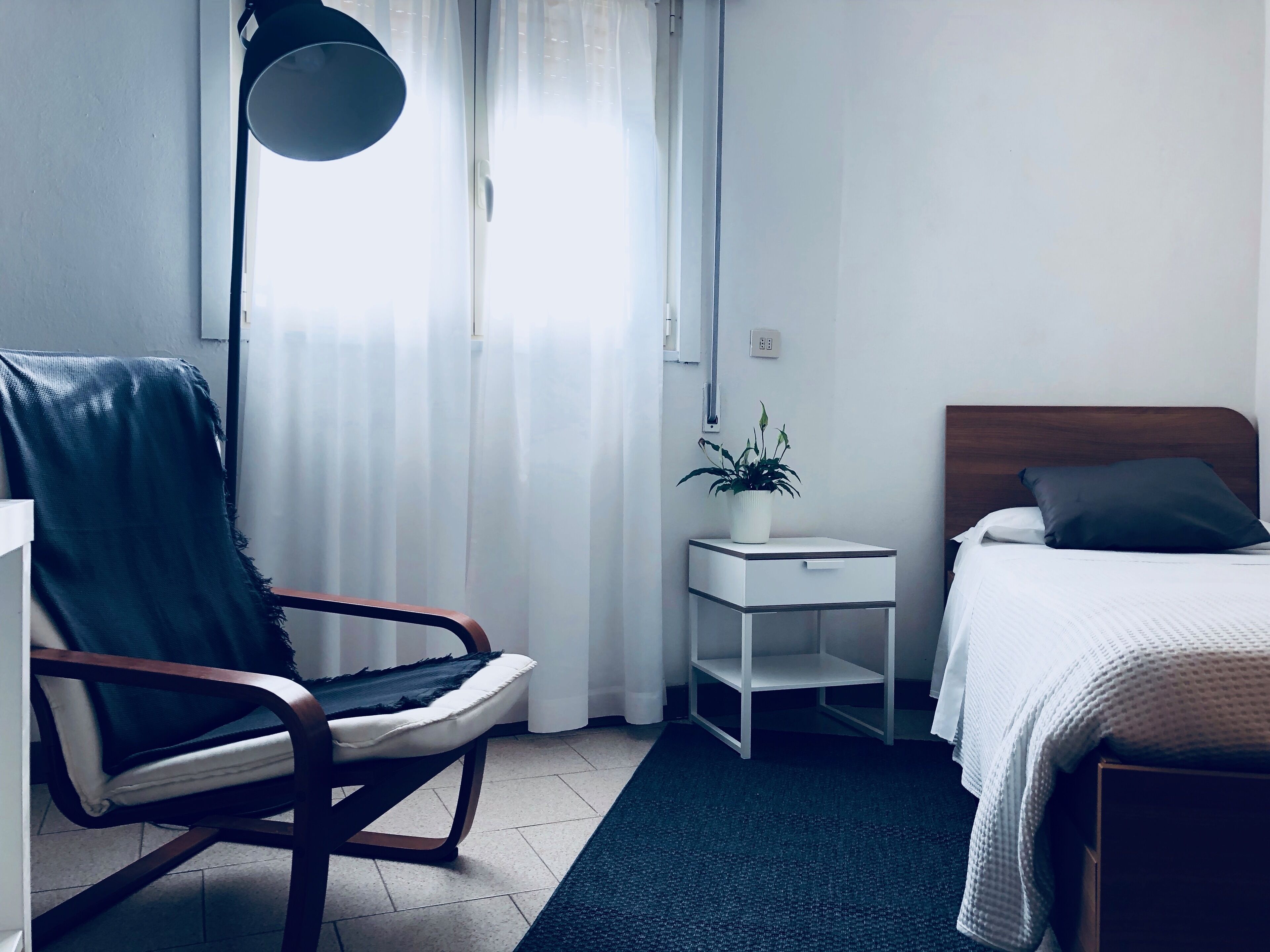 Une chambre d’hôtel avec un lit, une chaise, une table de chevet ornée d’une plante et une lampe sur pied.