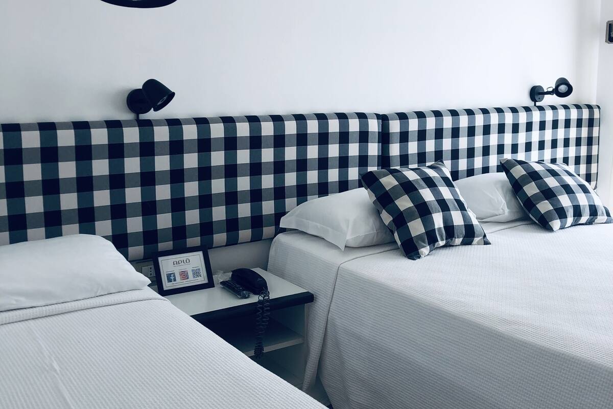 Habitación de hotel con cabecera a cuadros, dos camas, una mesita de noche con teléfono y una lámpara fijada a la pared.