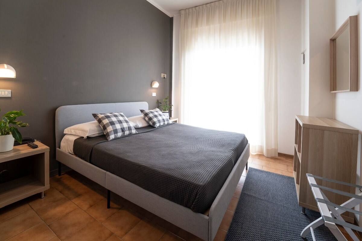 Un dormitorio moderno con una cama gris, almohadas estampadas, una mesita de noche con una planta y un teléfono, un mueble auxiliar de madera y un escritorio con una silla.