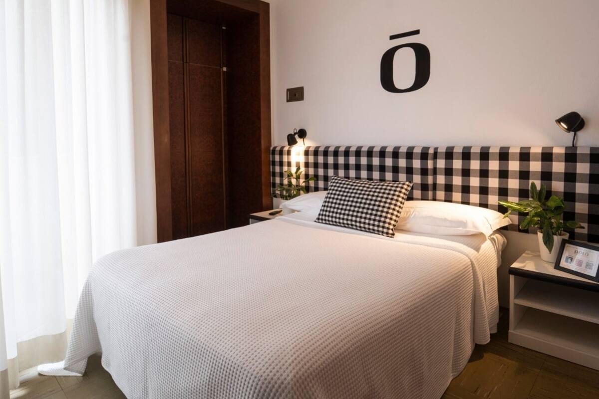 Un dormitorio ordenado con una cama grande, cabecera a cuadros, mesita de noche con lámpara y un aplique de pared.