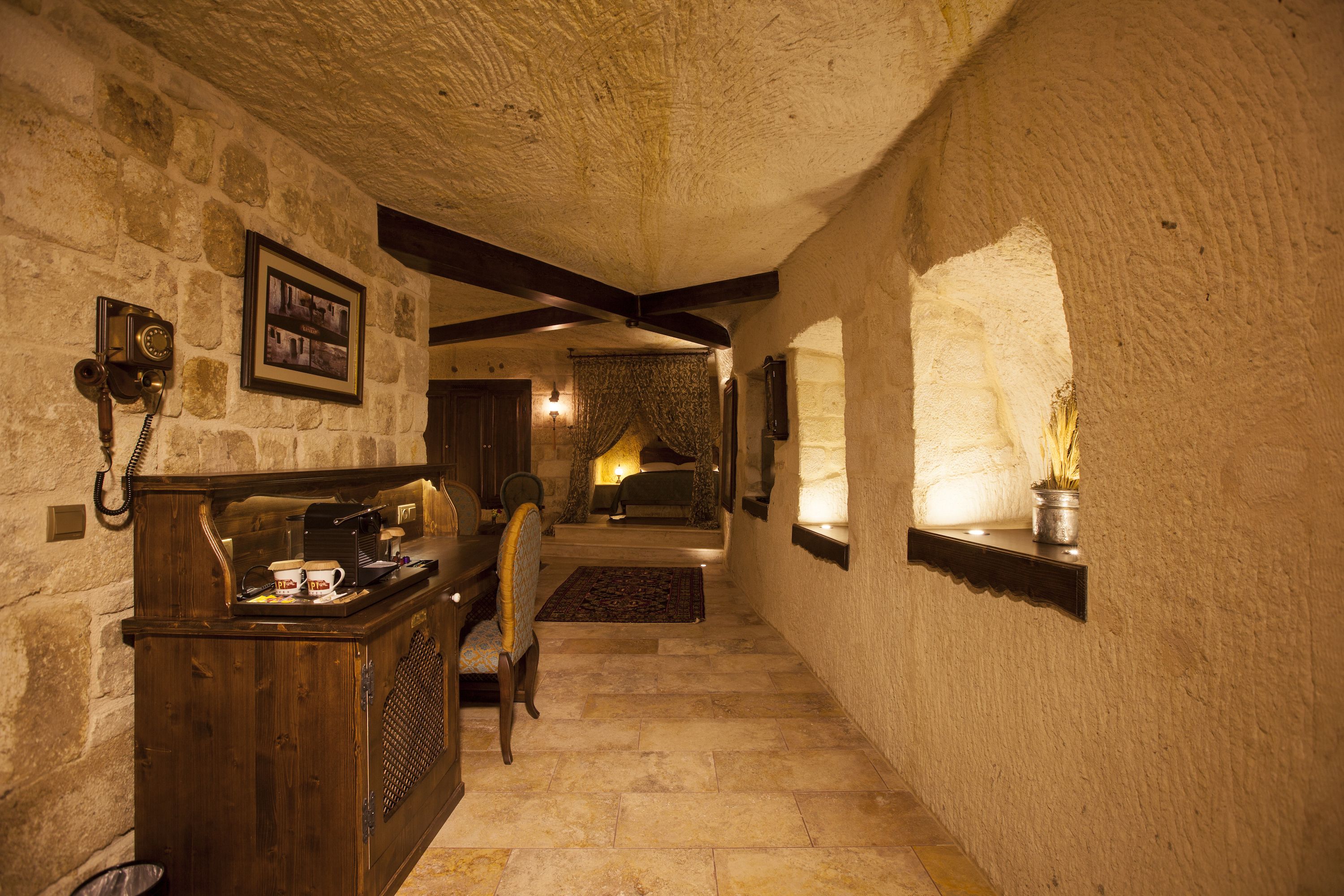 A narrow stone corridor with a wooden reception desk, a framed photograph, and a bed in the background.