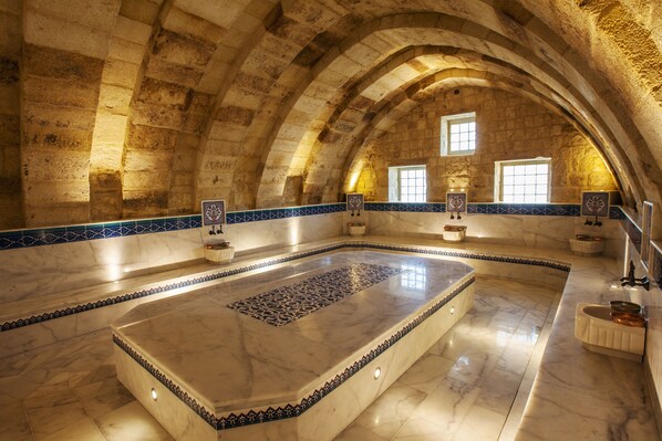 Una habitación de piedra en arco con una piscina central de mármol, paredes de azulejos y bancos de piedra.