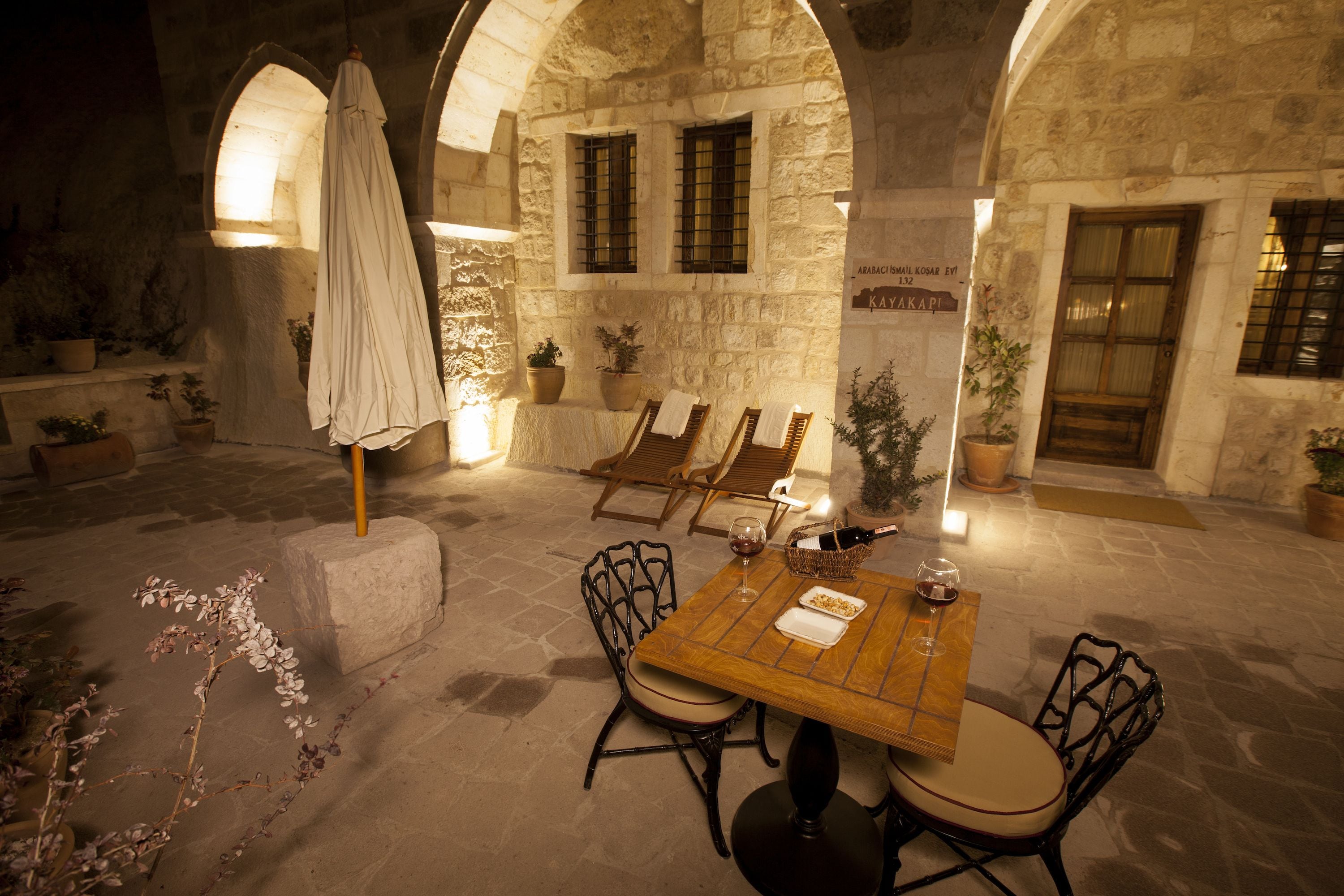 An outdoor dining area with stone walls, wooden tables, and chairs, surrounded by potted plants.