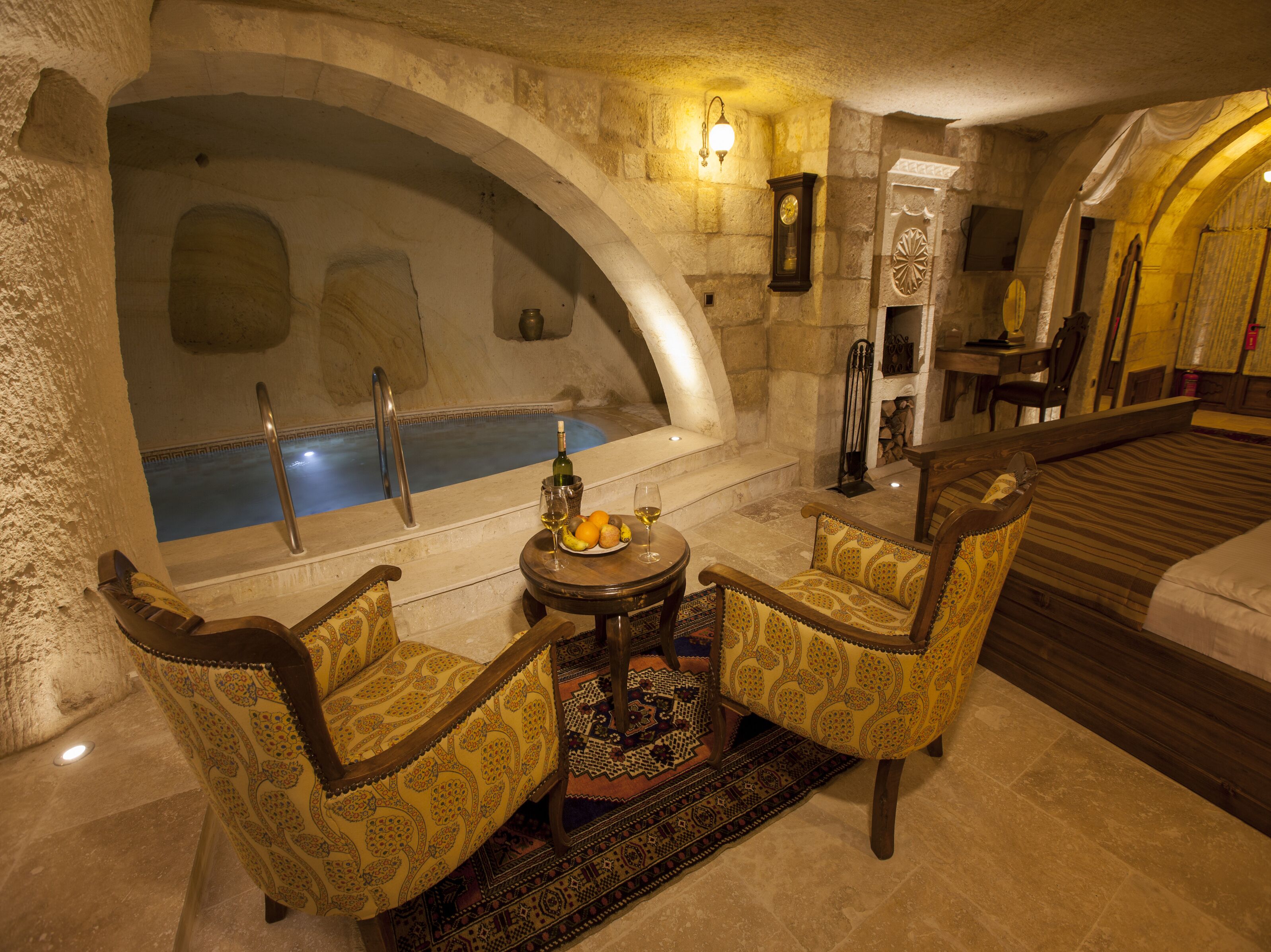 Una habitación con un arco de piedra, una bañera, dos sillones, una mesa con una botella y un vaso, y una cama.