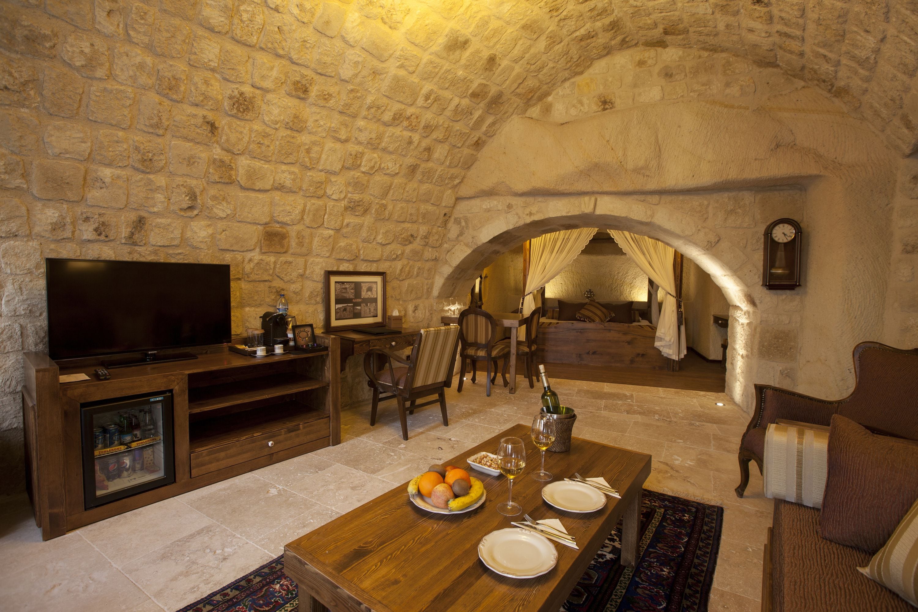 A stone-walled room with a television, wooden furniture, a dining table with plates and glasses, and a clock on the wall.