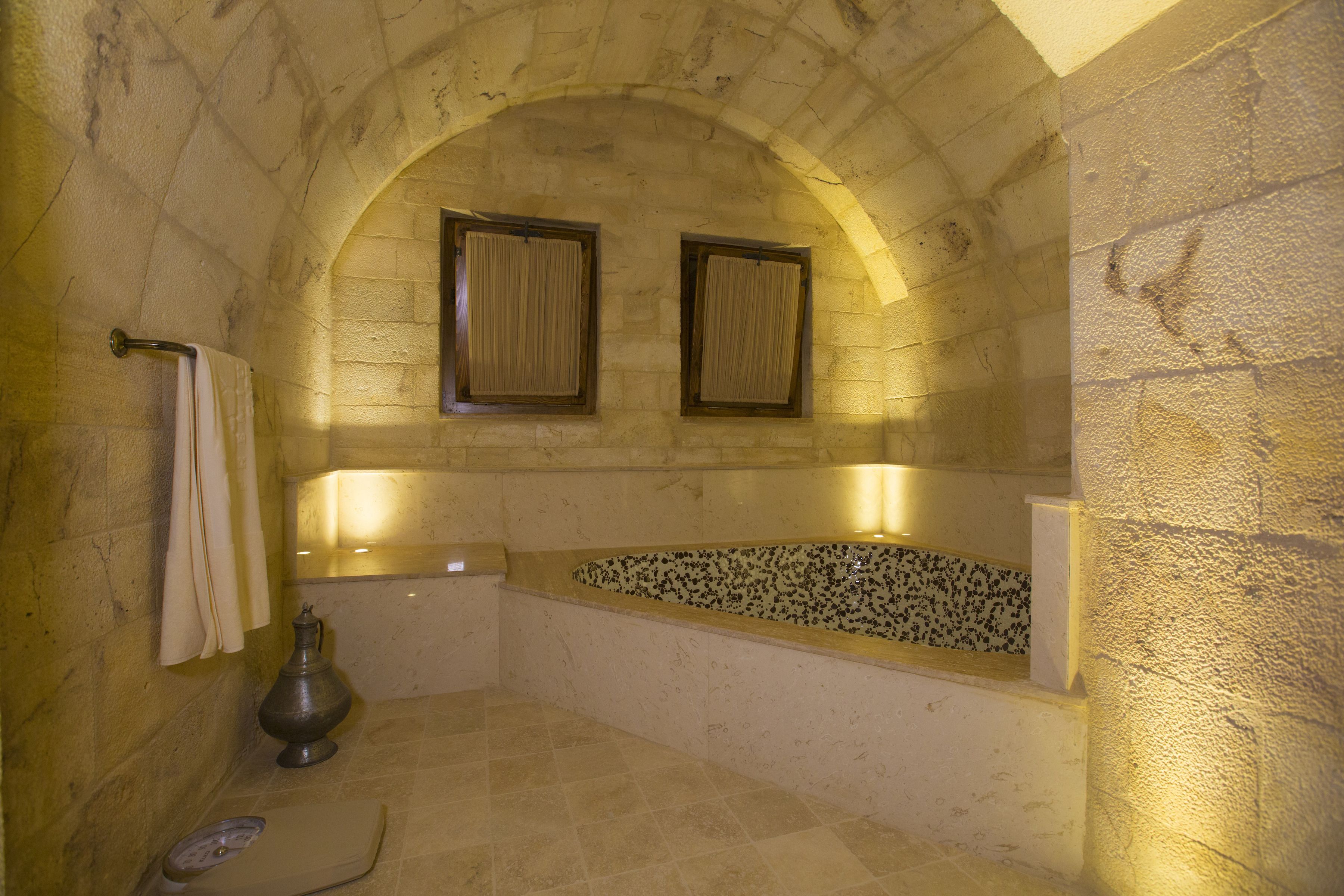 A stone-walled bathroom with a freestanding bathtub, a scale, and a metal container.