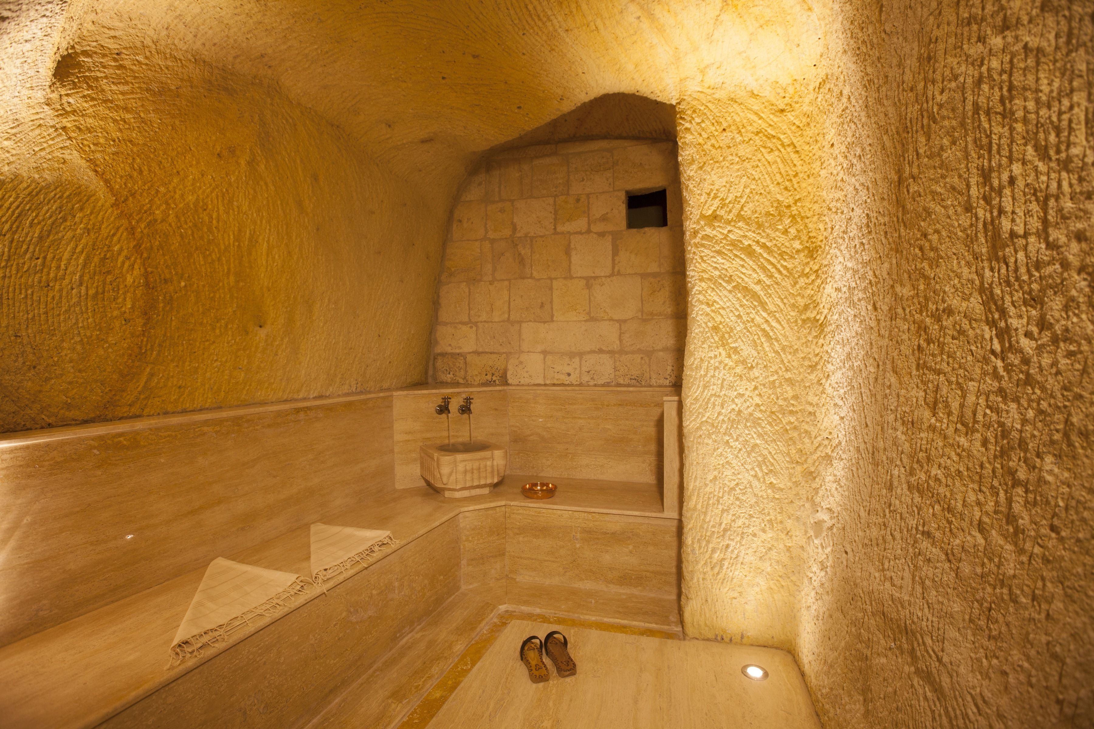A stone-walled, dimly lit interior with a bathtub, a small sink, and a pair of shoes on the floor.