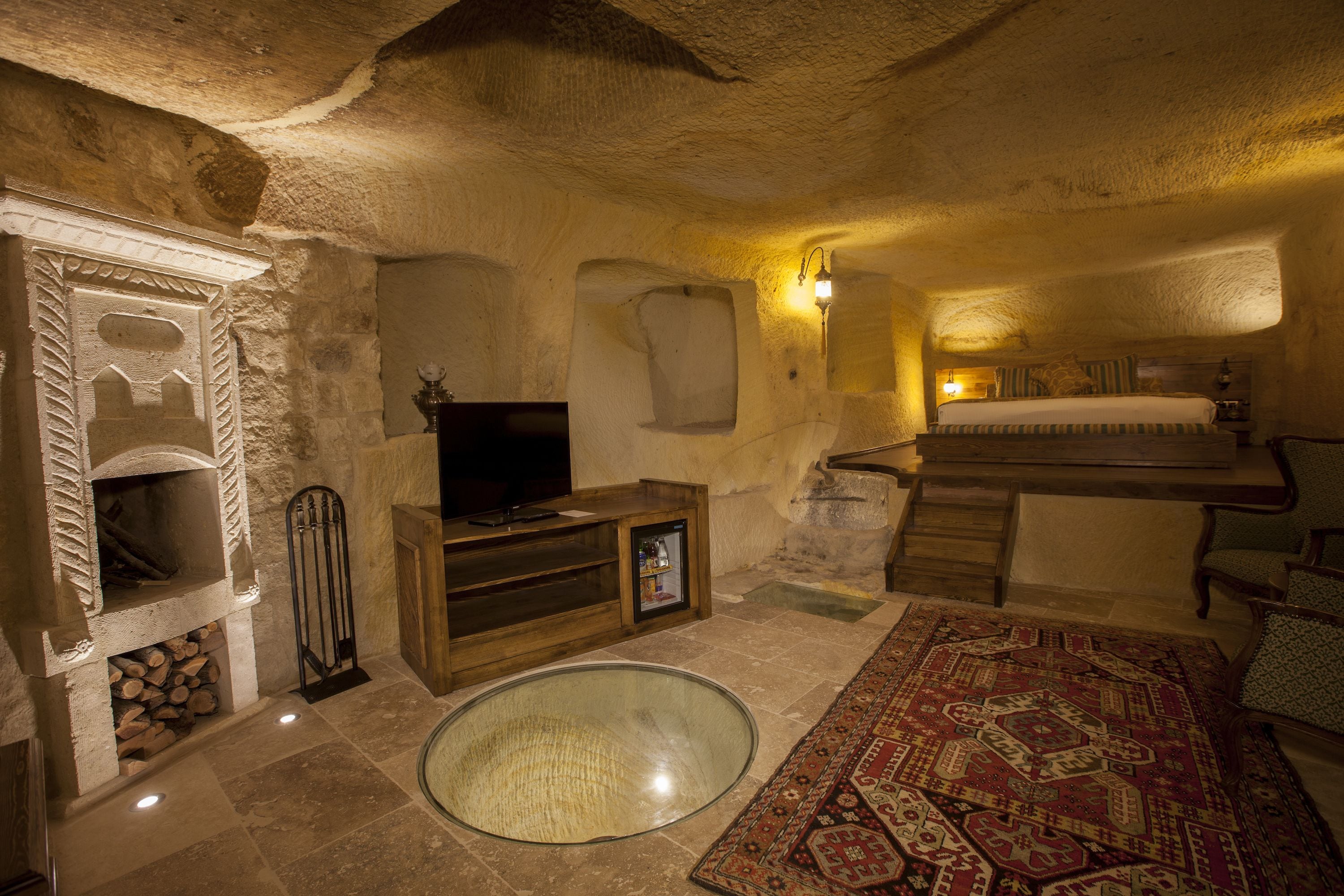 Una habitación de paredes de piedra con chimenea, televisor, refrigerador pequeño y cama con una alfombra estampada.