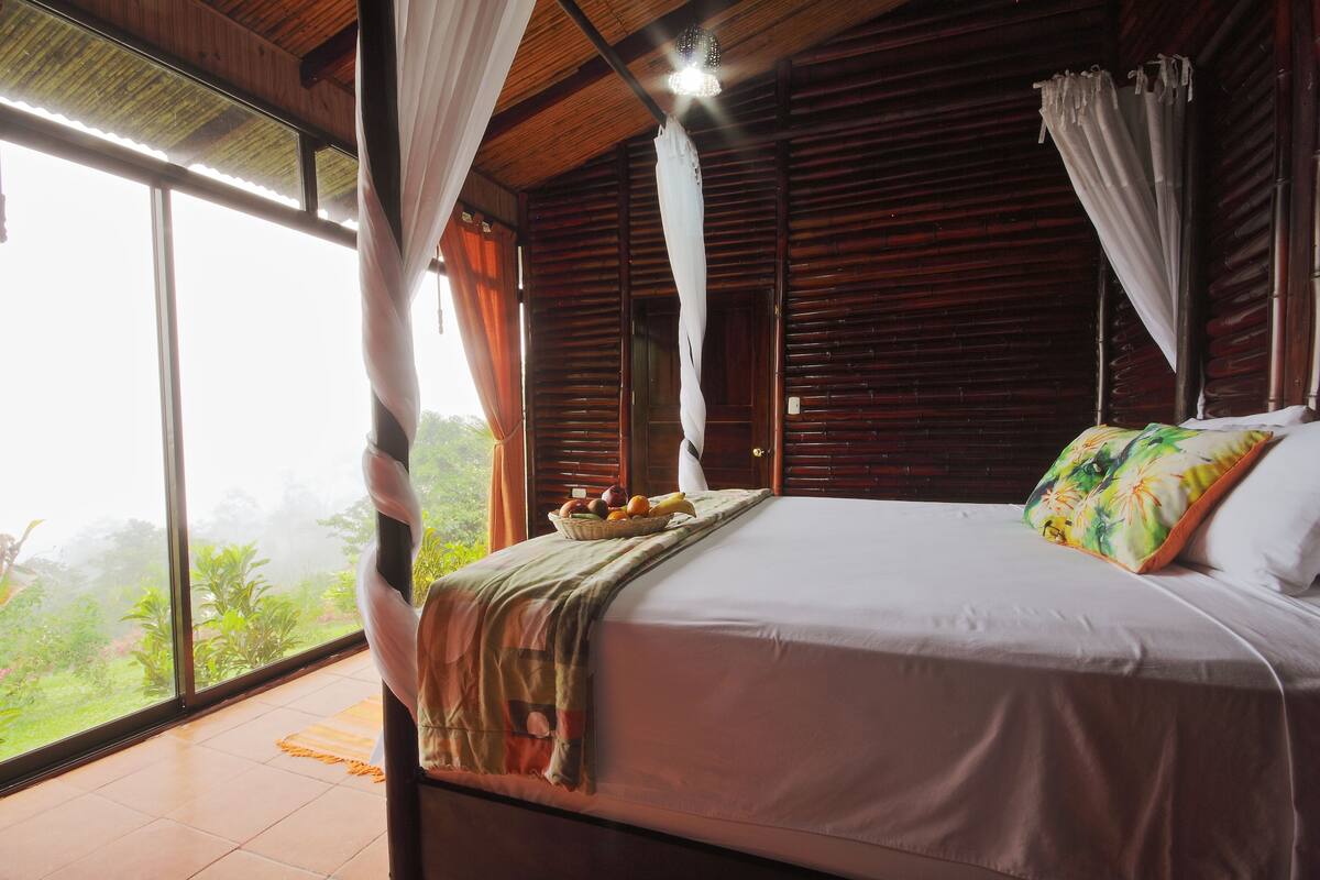 A bedroom with a large bed, a window with curtains, and a view of greenery outside.