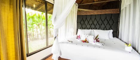 A large bed with white linens and a canopy, a dark headboard, and a view of greenery outside.