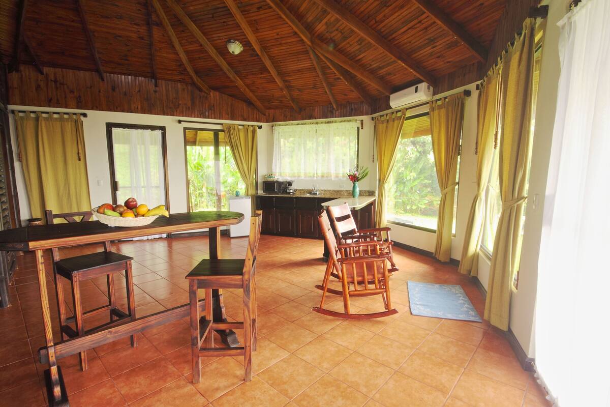 A spacious room with a wooden ceiling, a dining table with chairs, a rocking chair, and a kitchen area with a window.