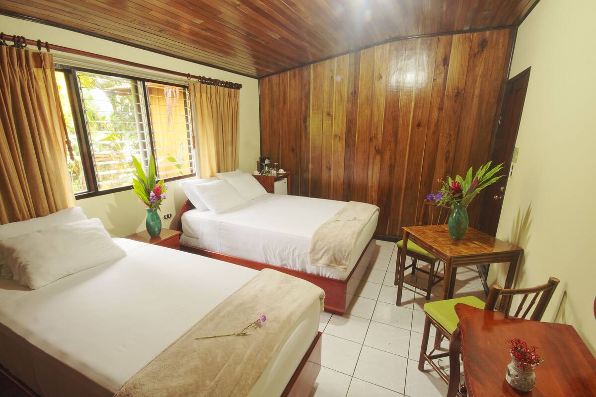 A hotel room with two beds, a wooden ceiling, a dining table with chairs, and a small table with a vase.