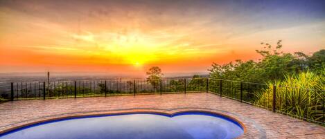 A swimming pool with a brick deck, surrounded by a metal fence, and a view of the sunset over a distant landscape.