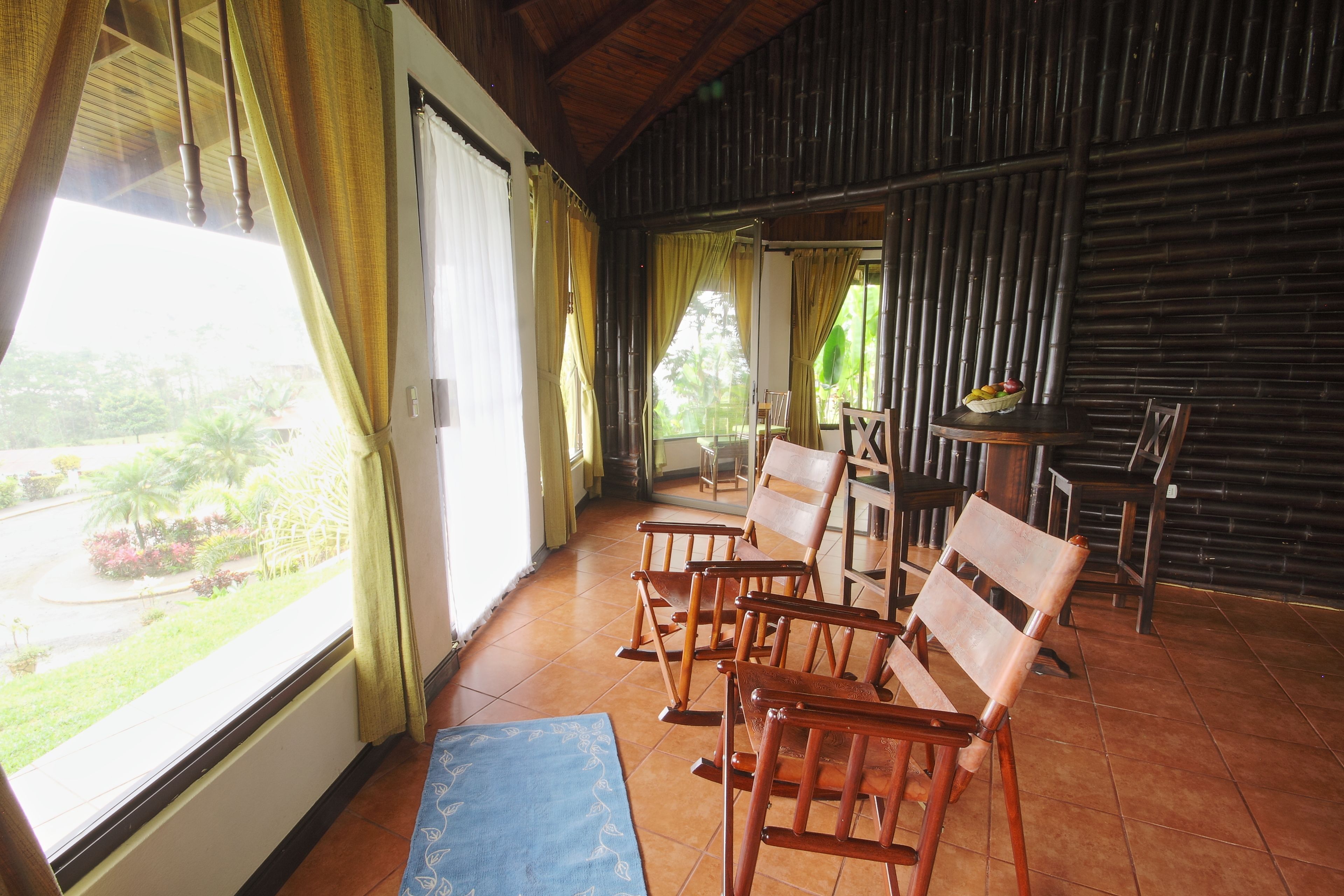 Una habitación con vista a un jardín, sillas de madera, una mesa y un tapete azul.