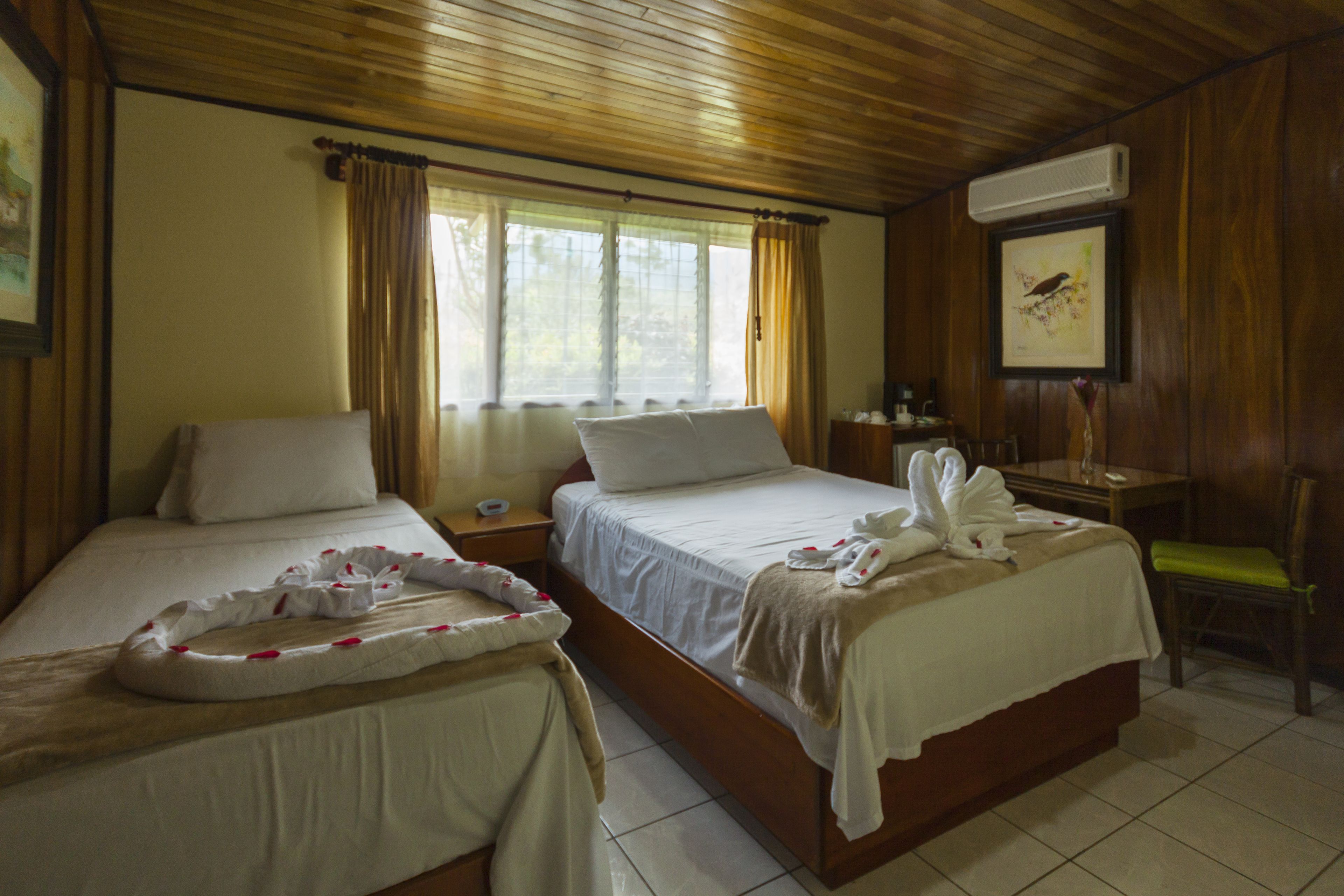 Habitación de hotel con dos camas, techo de madera, ventana con cortinas, escritorio con una silla y un cuadro en la pared.