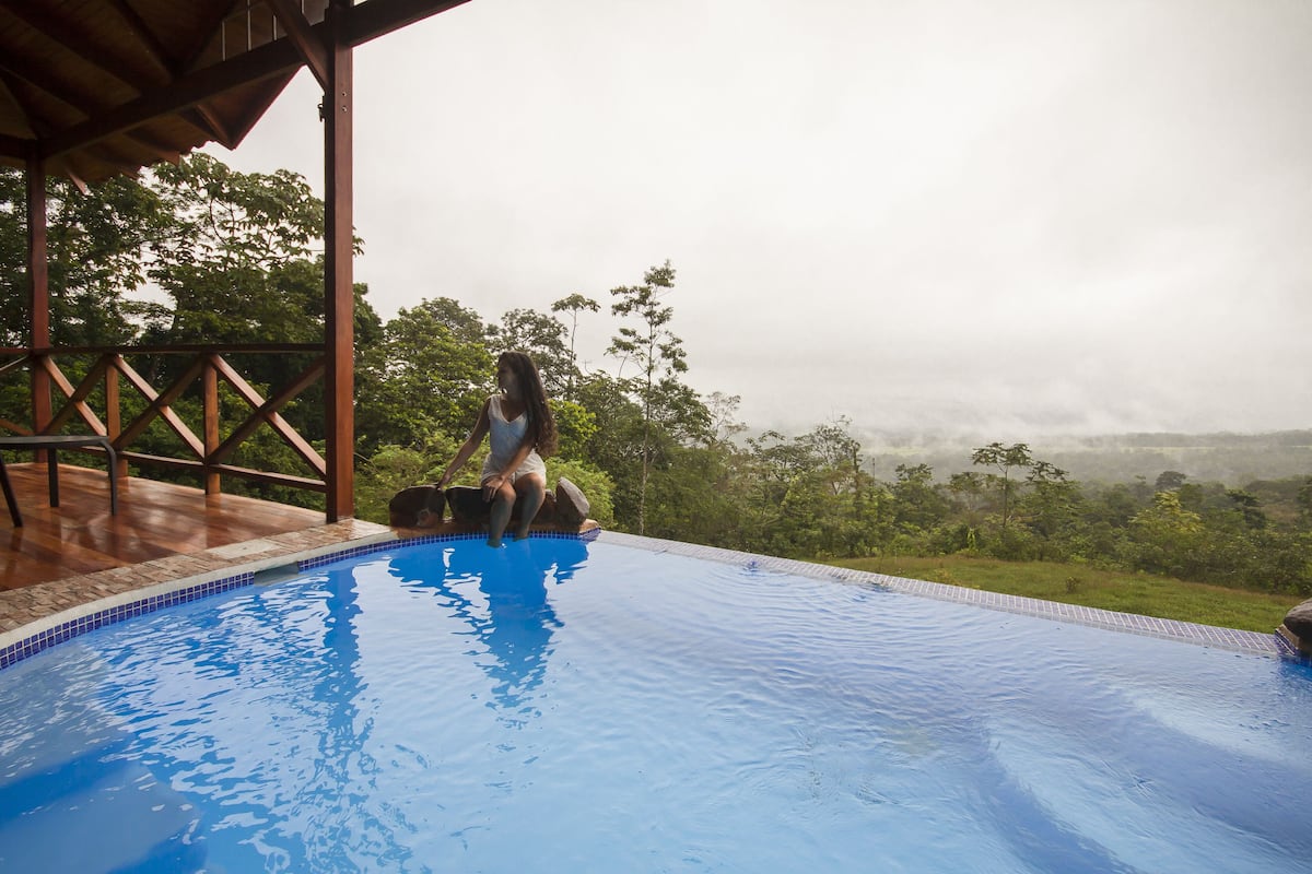 A woman sitting on a rock by a swimming pool with a view of a forested landscape.