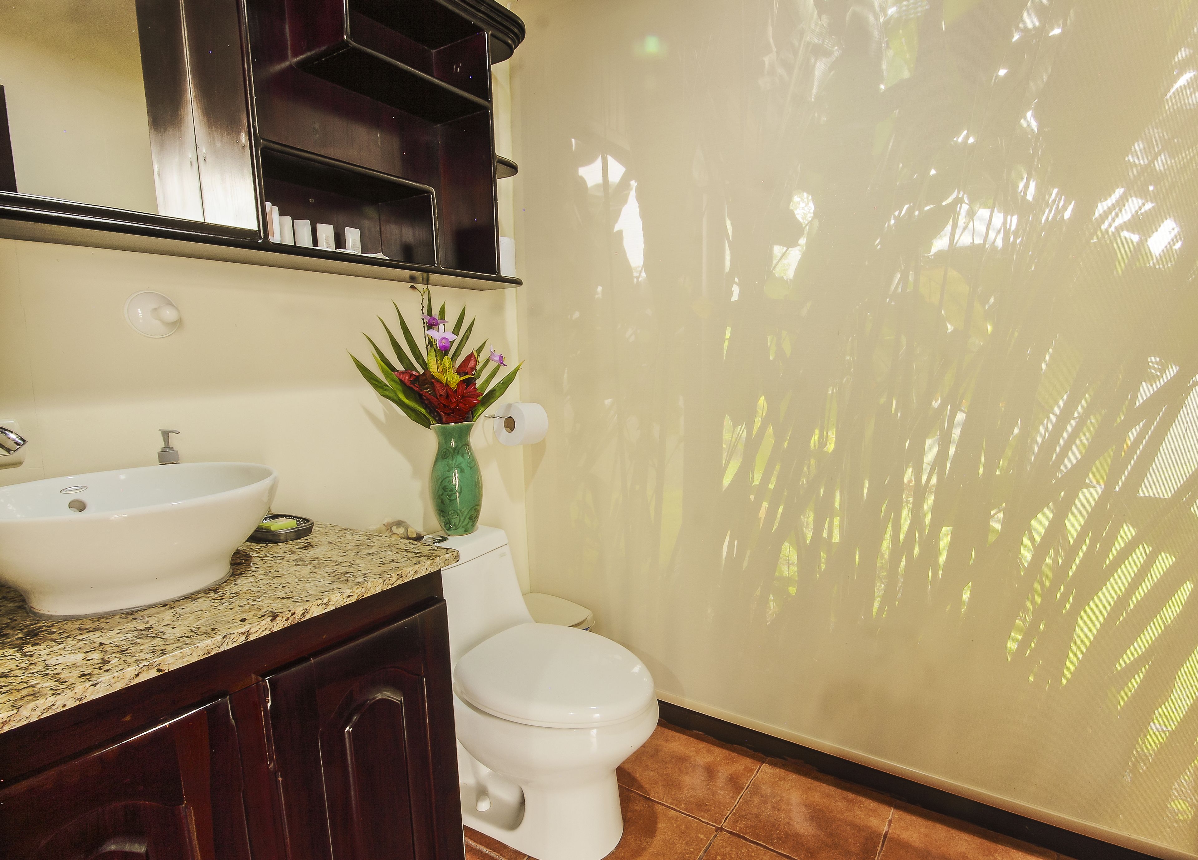 A bathroom with a white sink, a toilet, a green vase with flowers, and a mirror above the sink.