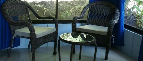 Dos sillas de mimbre con cojines y una mesita redonda en una habitación con vistas al exterior a través de una ventana.