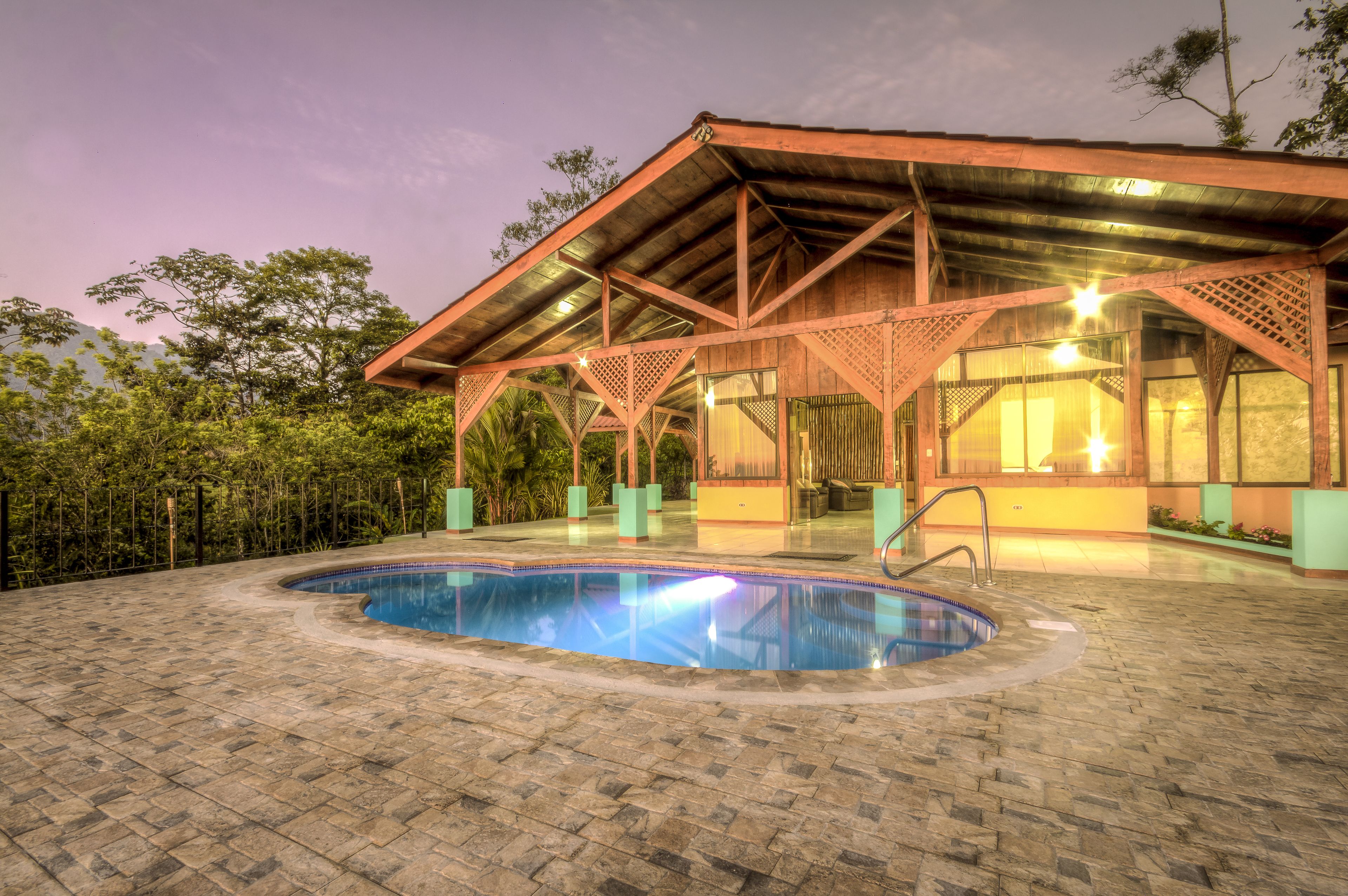 A pool area with a wooden structure overhead, surrounded by greenery.