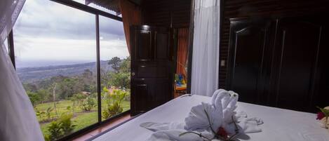 Un dormitorio con un ventanal grande, una cama con ropa de cama blanca y vistas a un paisaje verde.