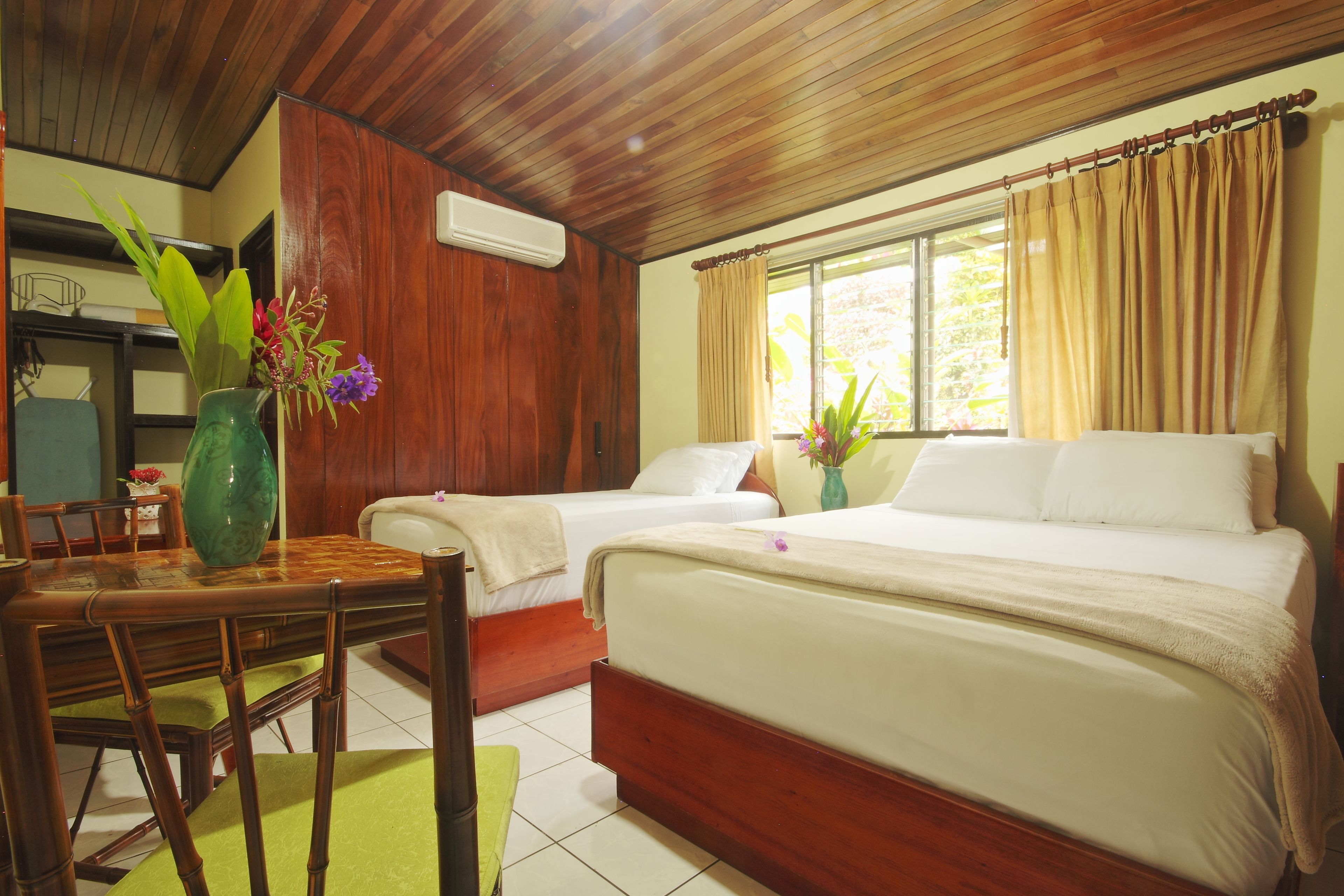 A hotel room with two beds, a wooden ceiling, a dining table with chairs, and a vase with flowers.