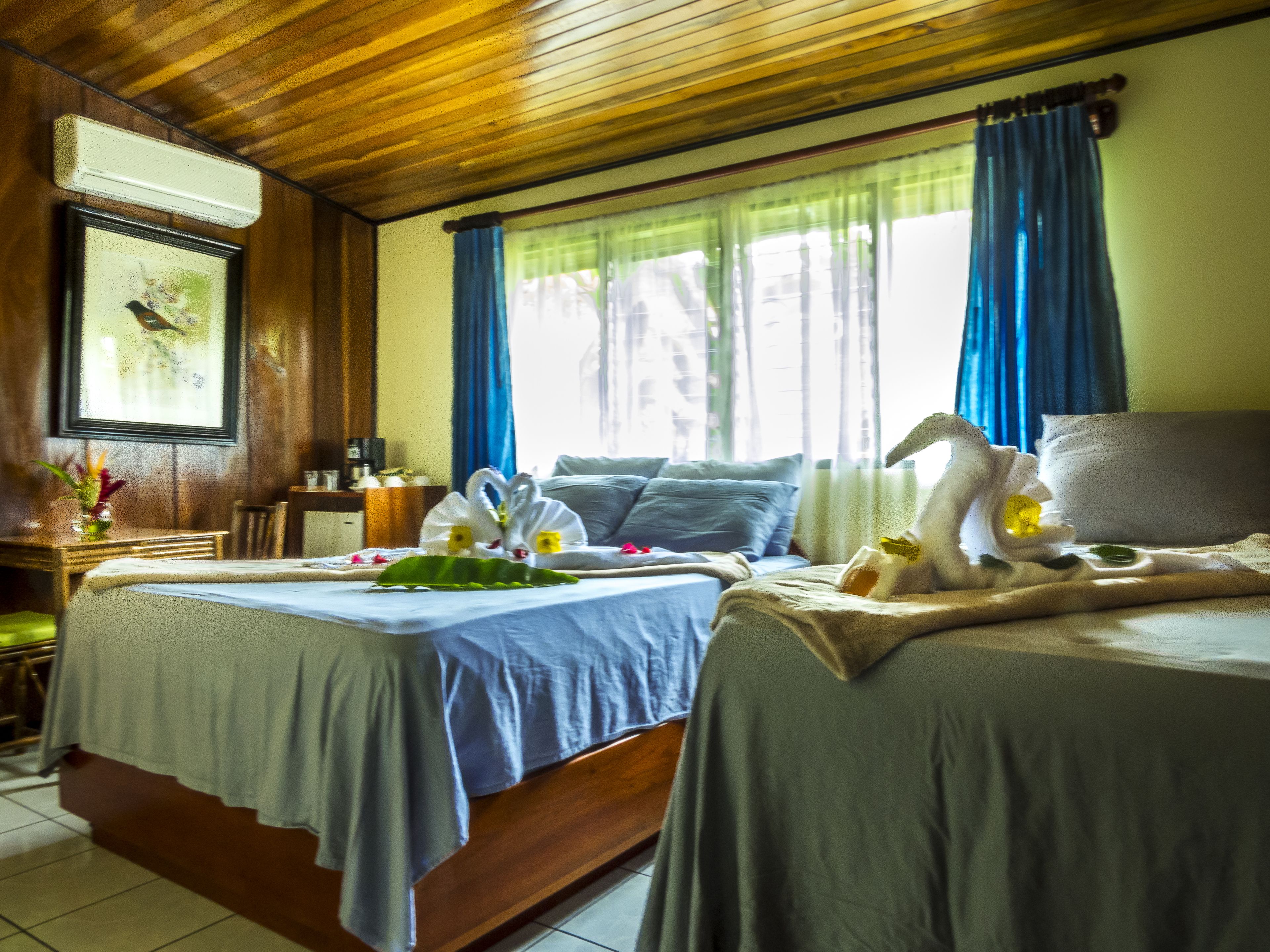 A hotel room with two beds, a wooden ceiling, a window with blue curtains, and a bedside table with a lamp.
