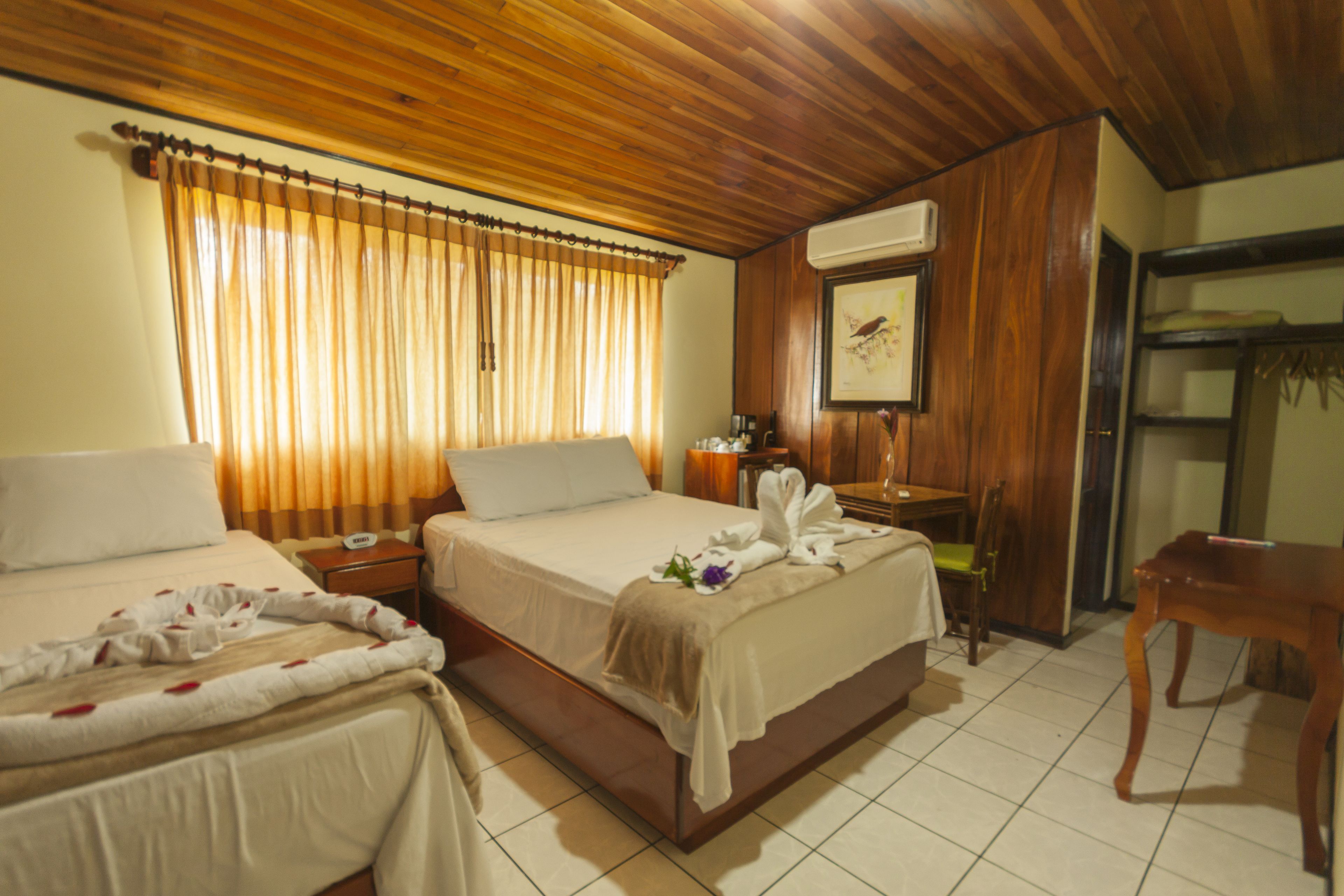 A hotel room with a wooden ceiling, a bed with a white comforter, a bedside table with a lamp, and a wooden chair.