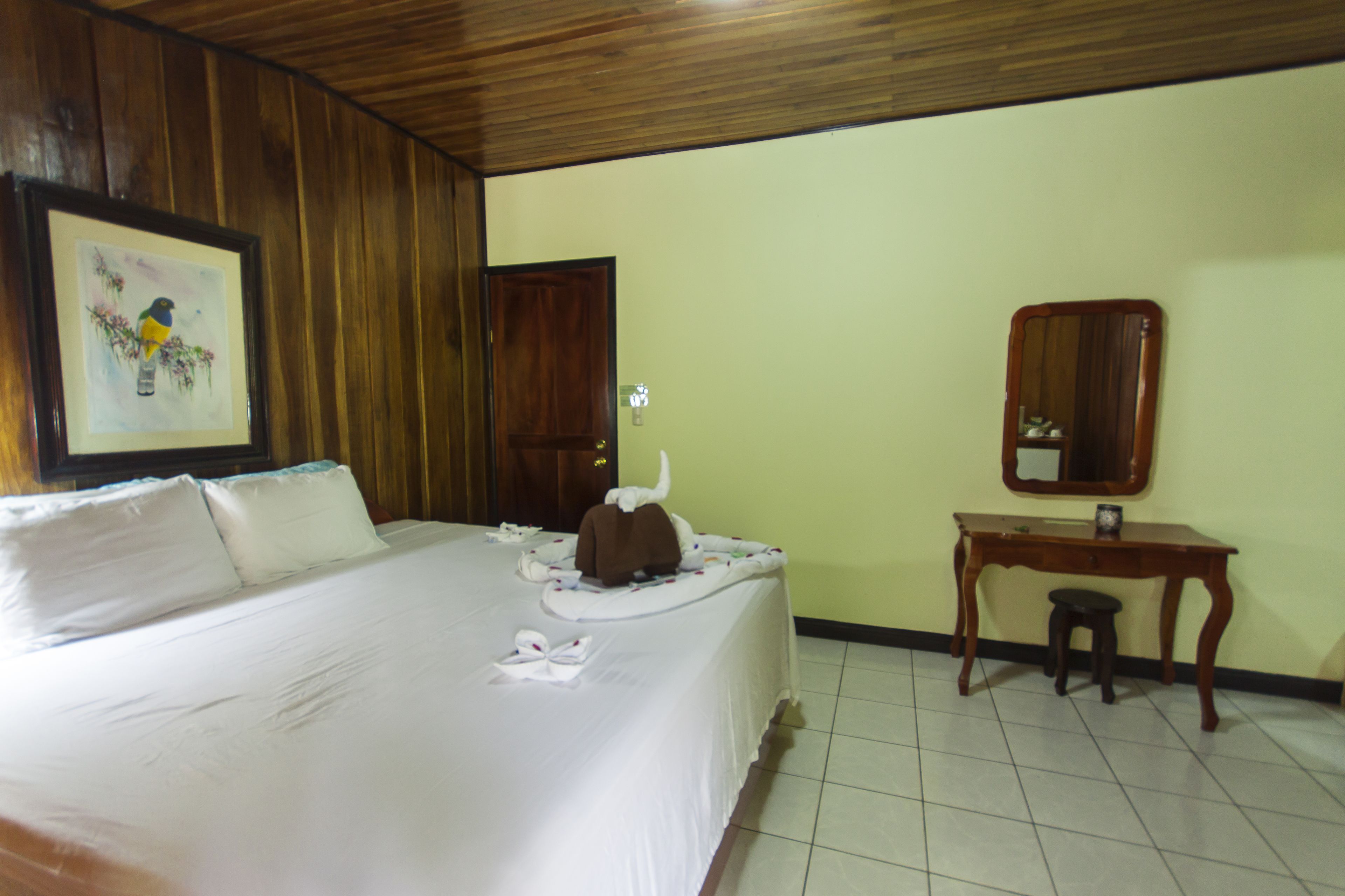 A hotel room with a large bed, a wooden headboard, a framed picture, a wooden desk, and a small stool.