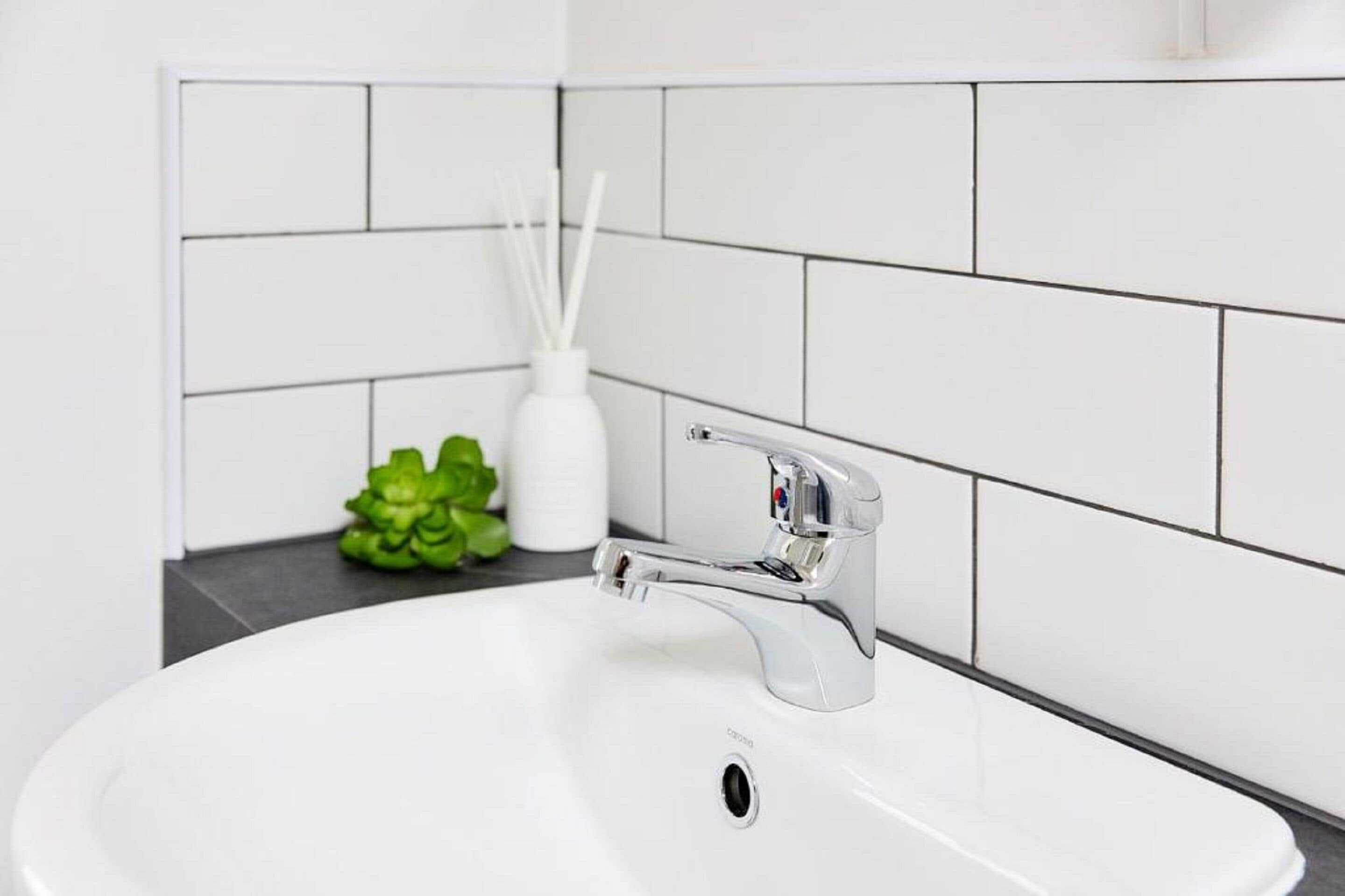 A white bathroom sink with a chrome faucet, white tiles on the wall, and a small green plant on the counter.