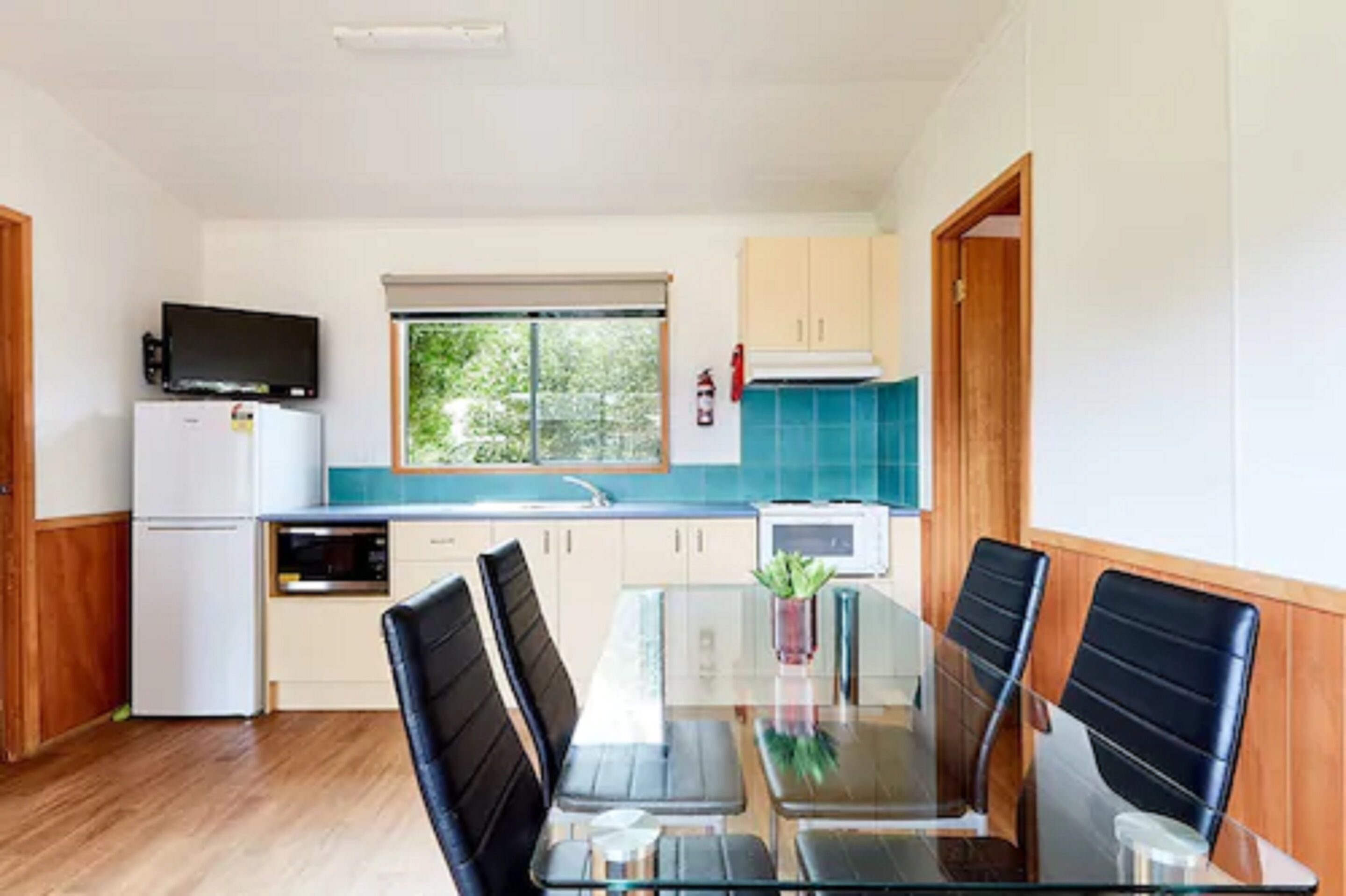 A modern kitchen with a dining area, featuring a glass table, black chairs, and a view of greenery outside.