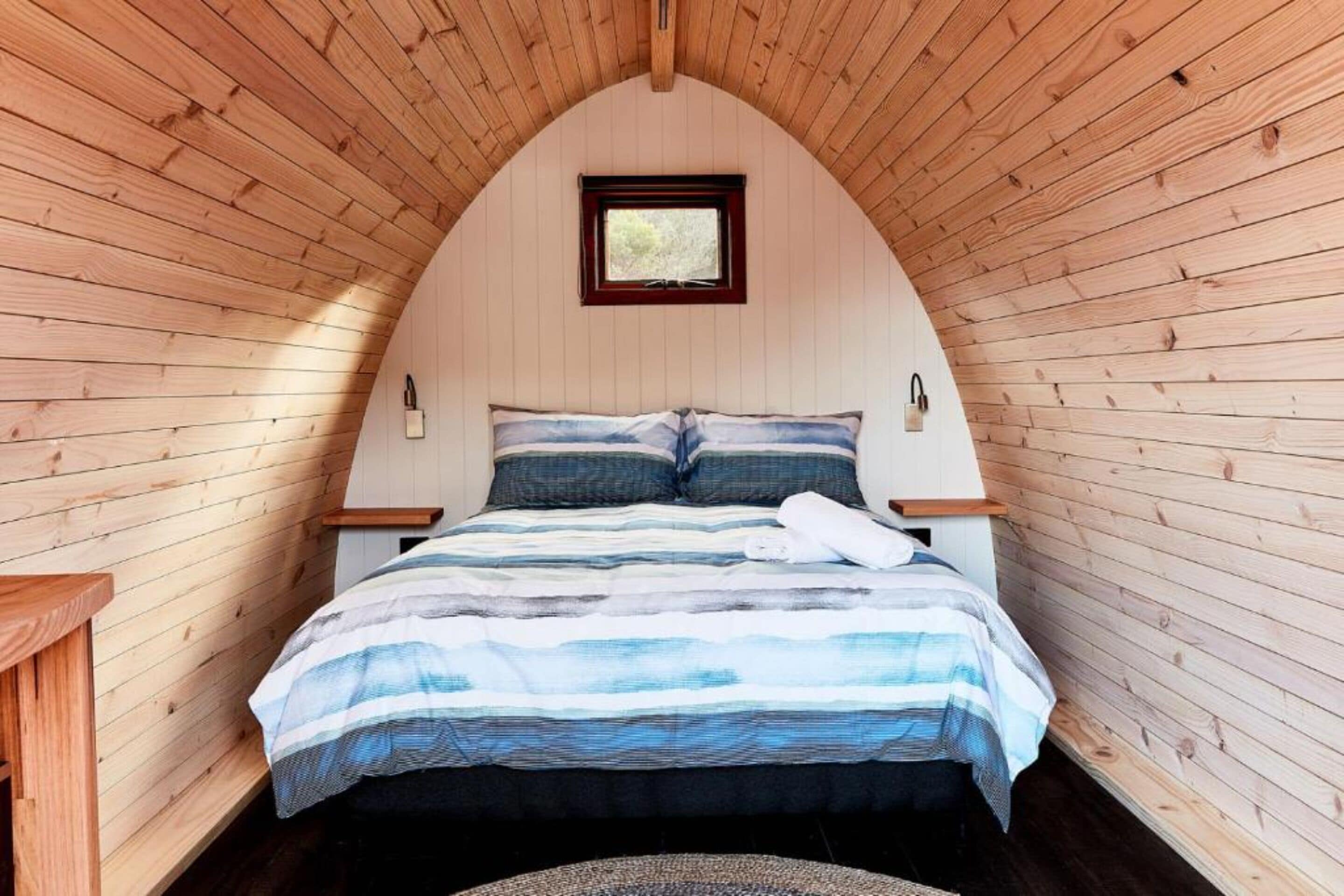 Une chambre confortable avec un plafond en pente en bois, un lit recouvert d’une courtepointe à rayures, une petite fenêtre et une table de chevet avec une lampe.