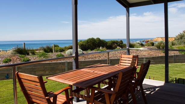 Un patio cubierto con una mesa y sillas de madera, con vistas a un paisaje costero con mar y vegetación.