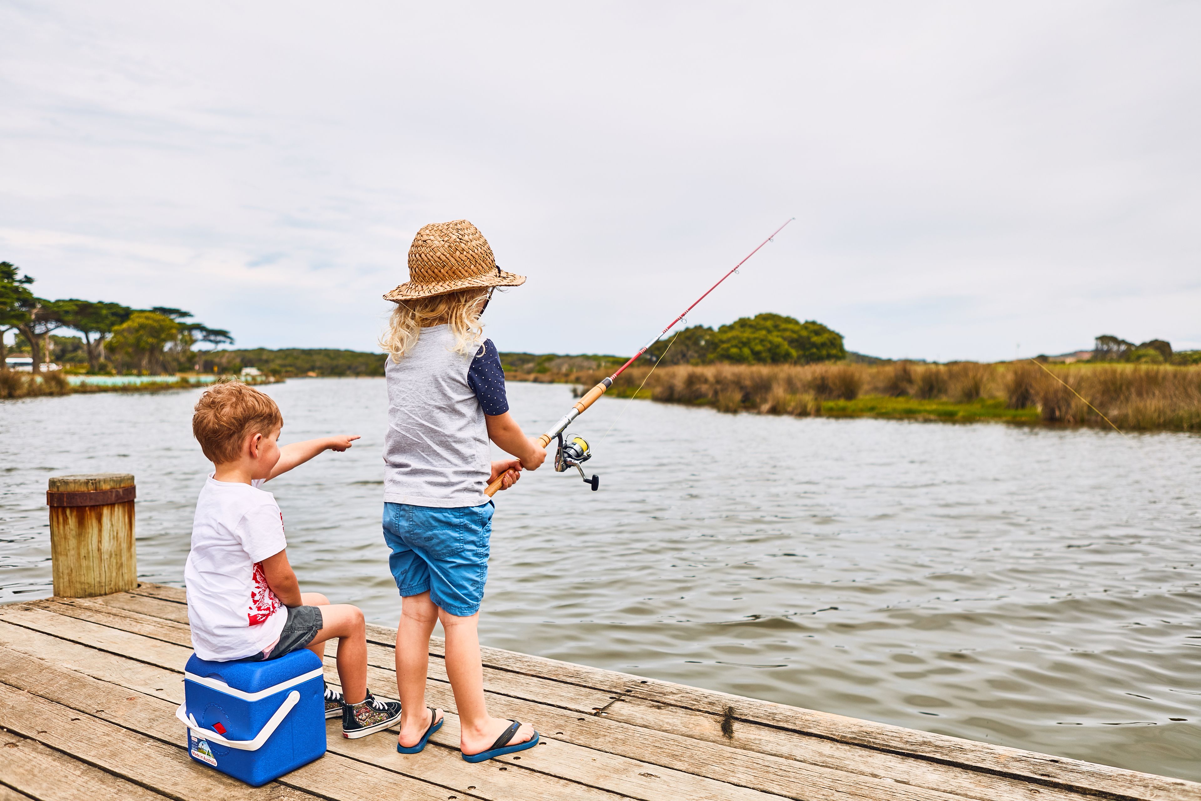Two people fishing on a wooden dock by a calm lake with trees in the background.