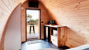 Una habitación compacta con paneles de madera, un escritorio integrado y un taburete pequeño, una puerta que da al exterior y un área exterior visible con vistas a árboles.