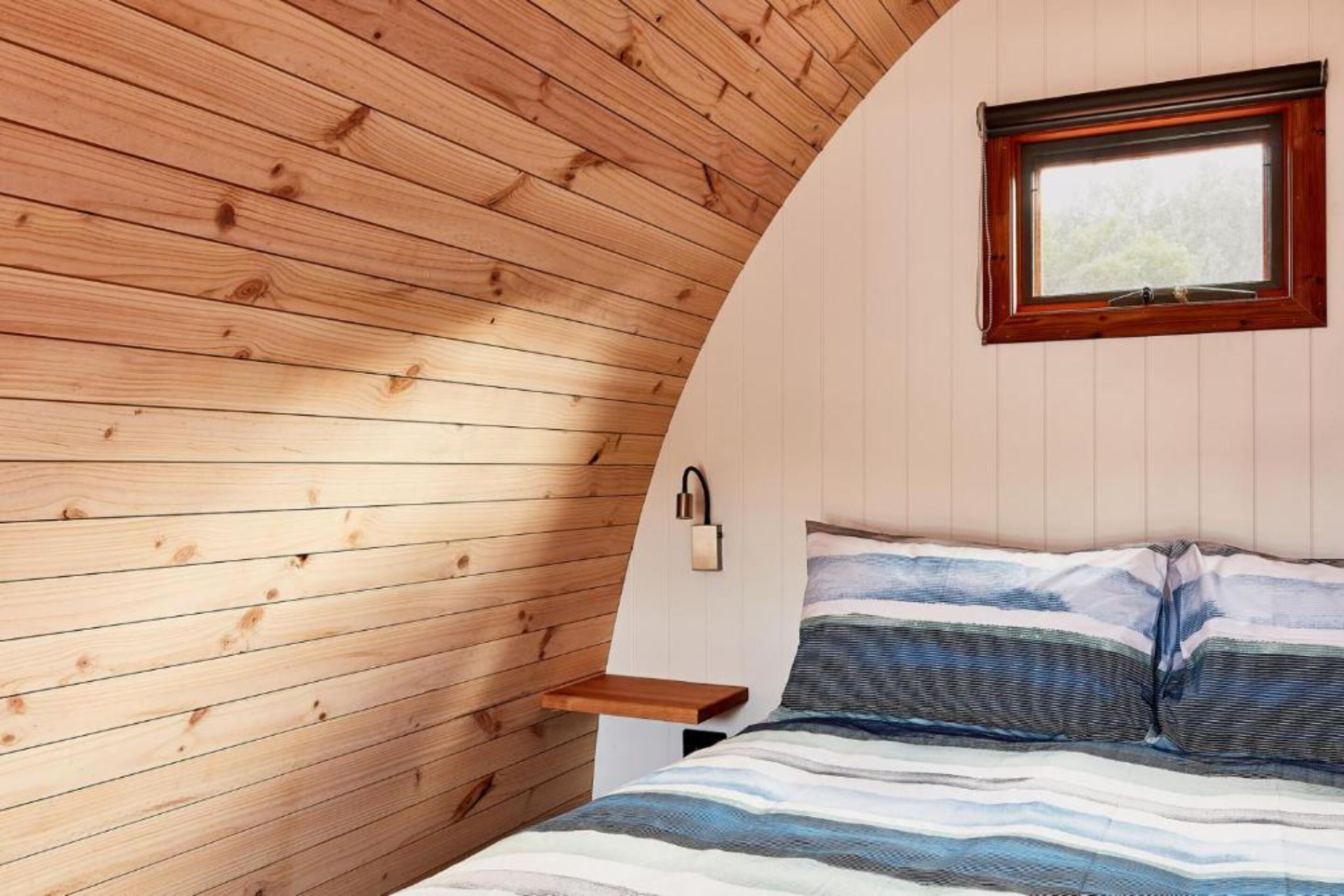 A bedroom with a wooden sloped ceiling, a bed with striped bedding, a nightstand, and a window with a view of trees.
