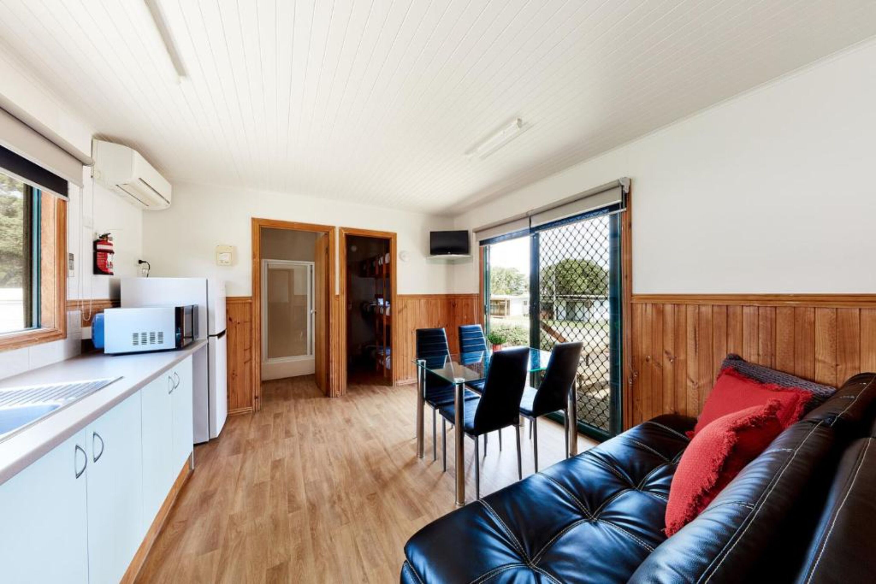 Et kompakt køkken med mikroovn, vask og brandslukker. Der er et spiseområde med bord og stole, samt en lædersofa med røde puder.